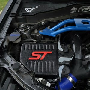 Könnte beinhalten: Motorraum eines Fahrzeugs mit schwarzem Luftfilterkasten mit rotem "ST"-Logo. Eine hellblaue Domstrebe und verschiedene Motorkomponenten sind sichtbar. Der Text "IMPROVED RACING" ist ebenfalls zu sehen.