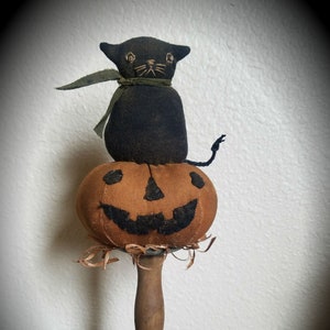 May include: A black cat with a green ribbon sits atop a brown pumpkin with a carved jack-o'-lantern face. The pumpkin is on a wooden stand with a gray base.  © T.F.C. Folk Art 2018