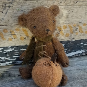 May include: A brown felt teddy bear wearing a green felt scarf and holding a brown felt pumpkin. The bear is sitting on a wooden surface. The text "T.F.C. Folk Art 2019" is visible in the image.