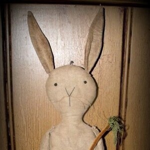 May include: A handmade primitive style bunny doll with long ears, brown pants, and a stick in its hand. The doll is made of fabric and has a rustic, vintage look.