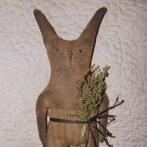 May include: A brown fabric bunny doll with long ears, wearing a brown fabric outfit and a black fabric belt. The doll is holding a bunch of dried flowers and twigs.