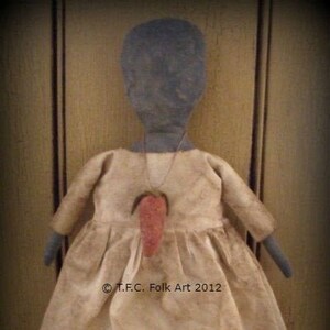 May include: A handmade rag doll with a black face, wearing a white dress with a red heart-shaped pendant. The doll is standing on a wooden surface. © T.F.C. Folk Art 2012