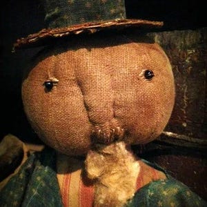 Pode incluir: Uma boneca de pano feita à mão com um rosto de estopa, olhos de botão pretos e uma barba de palha. A boneca está vestindo uma camisa listrada azul e branca com um colete listrado vermelho e branco e um chapéu estampado verde e branco.