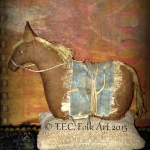 May include: A brown stuffed horse with a blue and white patterned saddle sits on a tan cushion. The horse has a long, flowing mane and tail. The cushion has the text "T.F.C. Folk Art 2015" printed on it.