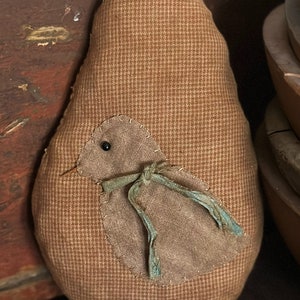 May include: A brown fabric bird with a blue ribbon, sewn onto a brown teardrop-shaped cushion. The cushion has a checkered pattern and the text (C) T.F.C. FOLK ART 2024 is visible on the bottom.