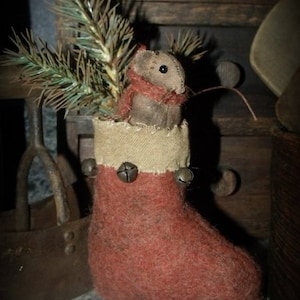 May include: A handmade felt stocking with a small, brown felt mouse peeking out of the top. The stocking is red with a white cuff and has jingle bells attached. The stocking is sitting on a wooden surface with a sprig of green pine.