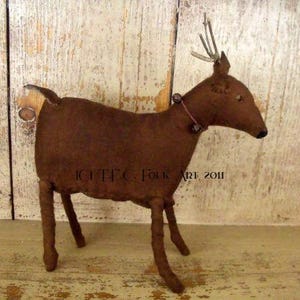 May include: A brown primitive folk art reindeer with a pink ribbon and bells around its neck. The reindeer has a long, thin tail and long, thin legs. The reindeer is standing on a wooden surface.