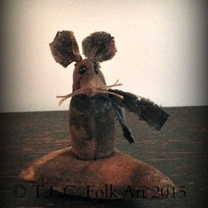 Pode incluir: Um rato de tecido castanho e preto com uma cauda longa e um corpo pequeno e redondo. O rato está sentado numa superfície de madeira. O texto "T.F.C Folk Art 2015" é visível no canto inferior direito da imagem.