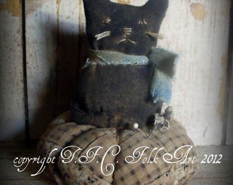 May include: Black fabric cat pincushion with a blue scarf, sitting on a brown and white checkered pincushion. The pincushion is filled with pins and needles. The text "copyright T.F.C. Folk Art 2012" is visible in the bottom left corner of the image.