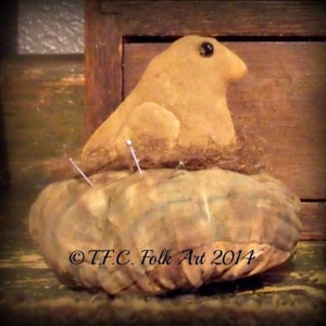 May include: A brown and tan fabric pincushion shaped like a bird's nest with a small bird sitting on top. The pincushion has several pins sticking out of it. The text "OTFC Folk Art 2014" is printed on the bottom of the pincushion.
