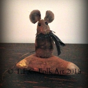 May include: A brown felt mouse with black button eyes and a black felt scarf is standing on a brown felt base. The base has a gold accent and the text "T.F.C. Folk Art 2015" is printed on the base.