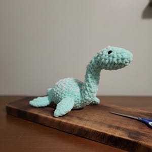 May include: A crocheted Nessie plush toy in light blue with pink accents. The handmade Loch Ness Monster has a long neck, small flippers, and a textured surface. The toy is resting on a wooden cutting board.