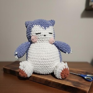 May include: A handmade crochet plush toy in the shape of a sleeping creature. The toy features a blue head and limbs, a white body, and brown accents on the feet. It has closed eyes and pink cheeks, resting on a wooden cutting board.