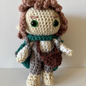 May include: A handmade crochet doll with a whimsical design. The doll features a cream-colored face, green eyes, and brown curly hair. It wears a green cape, gray overalls, and a brown satchel. The doll is approximately 6 inches tall.