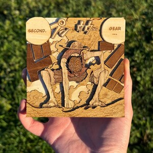 Peut inclure: Carré en bois avec un design découpé au laser représentant un personnage en chapeau de paille, accroupi. Les mots "SECOND" et "GEAR" sont inclus. L'œuvre est dans les tons de marron et de beige, avec des détails complexes.