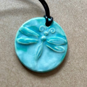 Könnte beinhalten: Ein türkisblauer Keramik-Anhänger mit Libellen-Design. Der Anhänger ist an einer schwarzen Schnur befestigt.
