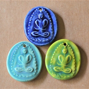 Puede incluir: Tres colgantes de cerámica con una figura estilizada en una postura de meditación sentada. Los colgantes son de forma ovalada y tienen un esmalte azul, verde y amarillo.