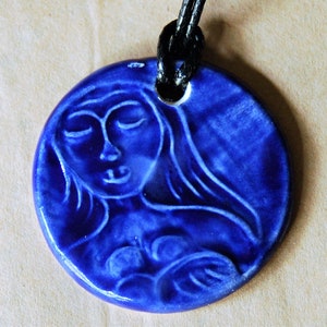 May include: A circular, deep blue ceramic pendant depicting a woman's face with closed eyes and long hair. The pendant is strung on a black cord. The woman's face is in relief, giving a 3D effect. This is a piece of handmade jewellery.