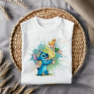 May include: White t-shirt with a cartoon illustration of Stitch holding a jar, with Tinkerbell flying nearby. The design features blue, yellow, and green accents, set against a burst of light. The shirt is displayed on a woven basket.