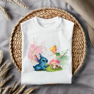 May include: White t-shirt with a blue Stitch character and a Tinkerbell character with pink wings. The characters are standing in front of a pink and green watercolor background with a mushroom.