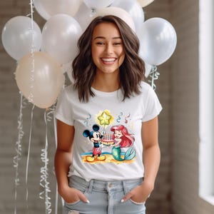 May include: White t-shirt with a colorful cartoon image of Mickey Mouse and Ariel from The Little Mermaid holding a birthday cake with candles. The image is surrounded by confetti and a star-shaped balloon.