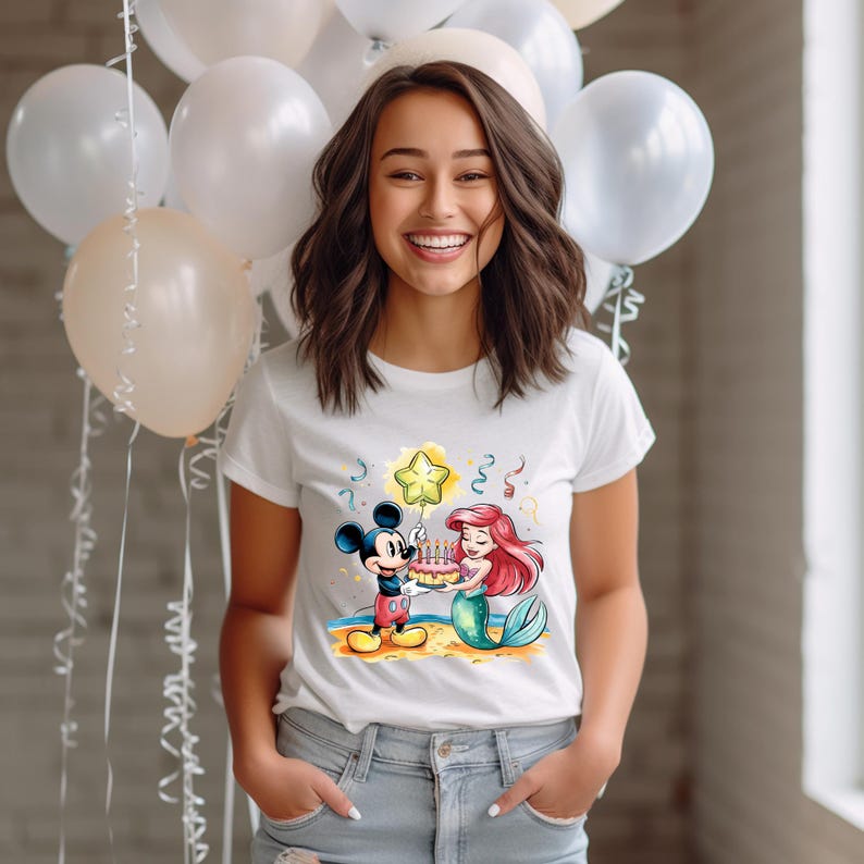 May include: White t-shirt with a colorful cartoon image of Mickey Mouse and Ariel from The Little Mermaid holding a birthday cake with candles. The image is surrounded by confetti and a star-shaped balloon.