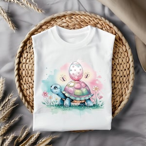 May include: A white t-shirt with a cartoon illustration of a turtle carrying an Easter egg. The turtle is blue and green, and the egg is pink and white with stars. The background is a watercolor wash of blue, pink, and green.