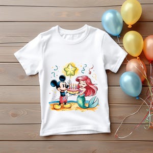 May include: White t-shirt with a colorful illustration of Mickey Mouse and Ariel the mermaid holding a birthday cake with candles. A yellow star is floating above them.