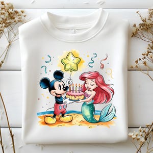 May include: A white sweatshirt with a colorful illustration of Mickey Mouse and Ariel from The Little Mermaid. Mickey is holding a pink birthday cake with lit candles and Ariel is holding the cake with a smile. A yellow star balloon is floating above them.