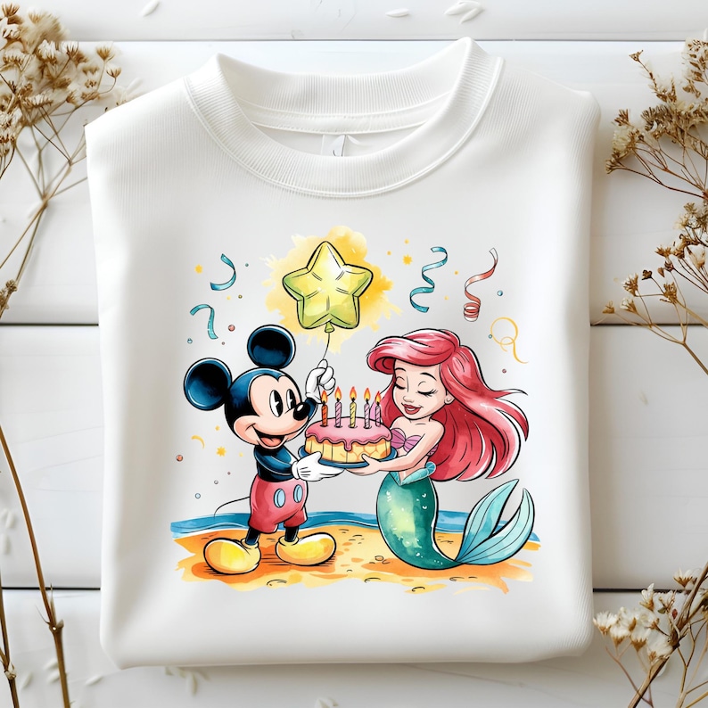 May include: A white sweatshirt with a colorful illustration of Mickey Mouse and Ariel from The Little Mermaid. Mickey is holding a pink birthday cake with lit candles and Ariel is holding the cake with a smile. A yellow star balloon is floating above them.