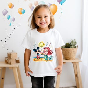May include: White t-shirt with a cartoon image of Mickey Mouse and Ariel the mermaid holding a birthday cake with candles. The image is surrounded by confetti and a star-shaped balloon.