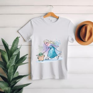May include: White t-shirt with a cartoon image of Elsa from Frozen ice skating with a Baby Yoda character.