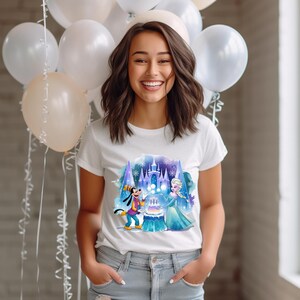 Może przedstawiać: Biały t-shirt z kolorową ilustracją zamku, Elsy z Krainy Lodu i Goofy'ego trzymającego tort urodzinowy. Ilustracja jest otoczona niebieskim i fioletowym efektem akwarelowym.