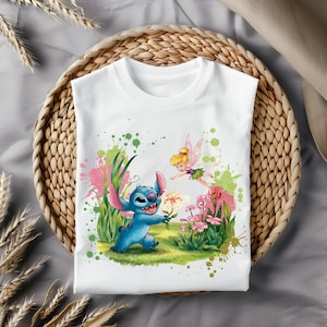 Puede incluir: Camiseta blanca con un personaje azul Stitch sosteniendo una flor y un personaje de Campanita volando sobre él. El diseño está rodeado de hierba verde, flores rosas y salpicaduras de pintura de colores.