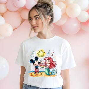 May include: White t-shirt with a colorful illustration of Mickey Mouse and Ariel from The Little Mermaid. Mickey is holding a birthday cake with candles and Ariel is holding a star-shaped balloon. The text "Happy Birthday" is printed on the shirt.