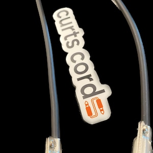 May include: Two black instrument patch cables with silver-colored right-angle metal plugs at both ends. Between the cables is a white rectangular sticker with rounded corners featuring the text curts cords in black and an orange icon of two plugs.