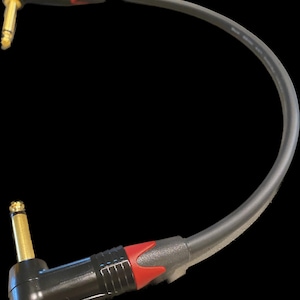 May include: A black audio patch cable featuring two right-angle 1/4-inch mono plugs. Each connector housing is black with a red accent ring and a gold-colored metal tip. The flexible black cable connects the two plugs in a curved shape.