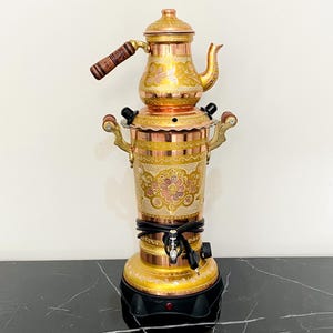 May include: A vintage copper and brass samovar with ornate floral engravings. The tea kettle has a wooden handle and sits atop a cylindrical base with a spigot. The samovar is gold and copper in colour and is displayed on a black marble surface.