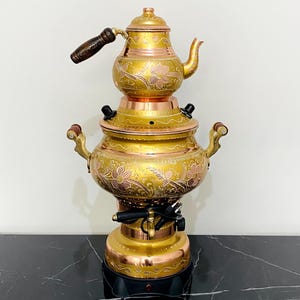 May include: A vintage brass samovar with detailed floral engravings. The tea kettle has a wooden handle and a decorative spout. The samovar has a copper base and a black electrical component. The design is ornate and traditional.