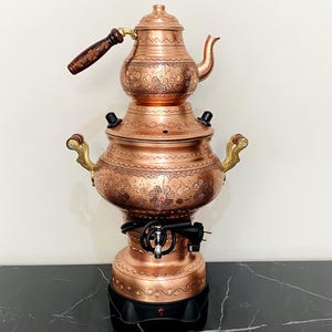 May include: A copper samovar with a teapot on top. The samovar features a floral pattern and brass handles. The teapot has a wooden handle. The samovar is on a black surface.