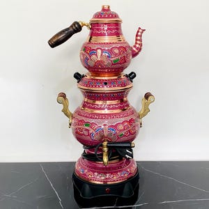 May include: An ornate red and gold samovar with a teapot. The samovar has a black base and gold handles. The teapot has a wooden handle. The design includes intricate patterns in blue, red, and gold. A traditional tea-making appliance.