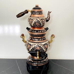 Pode incluir: Uma chaleira de chá de cobre e preta com desenhos florais ornamentados. A chaleira tem um bule em cima, uma câmara central e uma base com uma torneira. A alça e o bico são marrons escuros. A chaleira está em uma superfície de mármore preto.