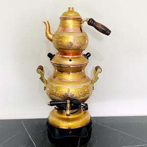 May include: An antique gold-coloured samovar with copper detailing. It features a teapot, a spigot, and brown handles. The base is black. The samovar is decorated with detailed patterns. The height is approximately 50 cm.