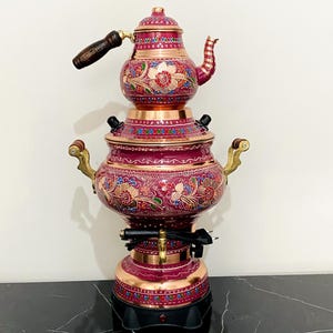 May include: An ornate samovar and teapot set. The samovar is burgundy with copper accents and floral designs. The teapot has a wooden handle. The base is black, and the samovar has brass handles. The height is approximately 50 cm.
