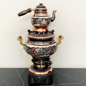 May include: A decorative copper samovar with detailed floral patterns and a black base. The tea kettle features a wooden handle and spout. The samovar is a traditional tea-making appliance, often used for brewing tea. The height is approximately 40 cm.