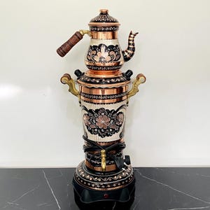 May include: A copper and black samovar with floral patterns. The tea kettle sits on a cylindrical base with ornate detailing. The samovar has a wooden handle and brass accents. The base is black with a decorative pattern.
