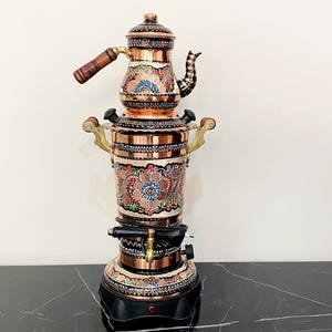 May include: A copper samovar with floral patterns in red, blue, and green. The samovar has a teapot on top with a wooden handle and a black base. The handles are gold-coloured. The samovar is on a black marble surface.