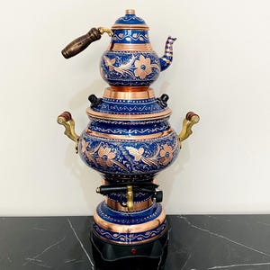 May include: A decorative copper and blue samovar with floral and bird designs. The tea kettle has a wooden handle and brass accents. The samovar is a traditional tea-making appliance.
