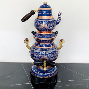 May include: A blue and copper Turkish samovar with detailed floral designs. The tea kettle features a wooden handle and brass elements. The samovar is a traditional tea-making appliance.