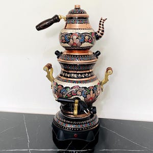 May include: An ornate copper and black electric samovar, featuring floral and geometric designs. The teapot has a wooden handle and a decorative spout. The base is black with a control panel and a spigot. A traditional tea-making appliance.